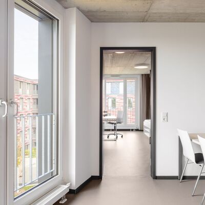 Blick in ein m�bliertes Zimmer des neuen Studierendenwohnheims Westside am 22.10.2026. Das Zimmer ist hell erleuchtet durch ein gro�es Fenster, das den Blick nach drau�en freigibt. Neben dem Fenster befindet sich eine T�r, die in einen weiteren Raum f�hrt. Das Zimmer ist modern eingerichtet mit einem Tisch, vier wei�en St�hlen und einem Schreibtisch im Hintergrund. Die W�nde sind wei�, die Decke aus Beton, und der Boden mit einem abget�nten glatten Belag ausgelegt.