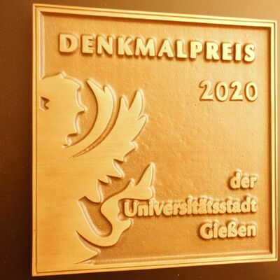 Goldene Plakette mit der Inschrift "Denkmalpreis 2020 der Universitätsstadt Gießen" und einer Prägung eines Teils des Gießener Wappentiers.