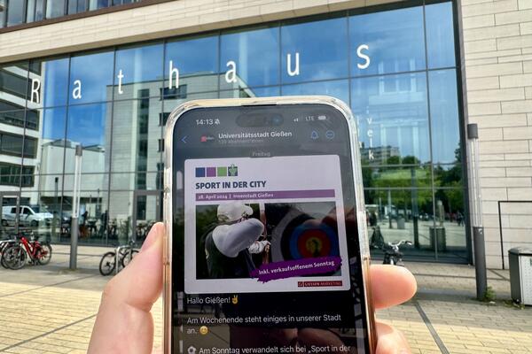 Ein Smartphone mit aufgerufenem WhatsApp-Kanal der Stadt Gie�en, im Hintergrund die Glasfassade des Rathauses